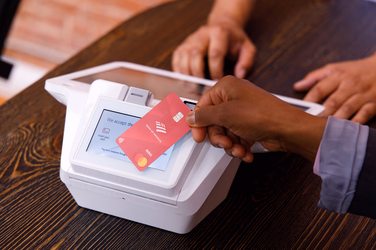 A hand tapping a contactless card against a payment terminal to pay at a counter