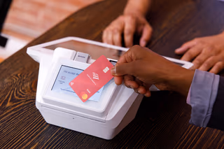 A hand tapping a contactless card against a payment terminal to pay at a counter