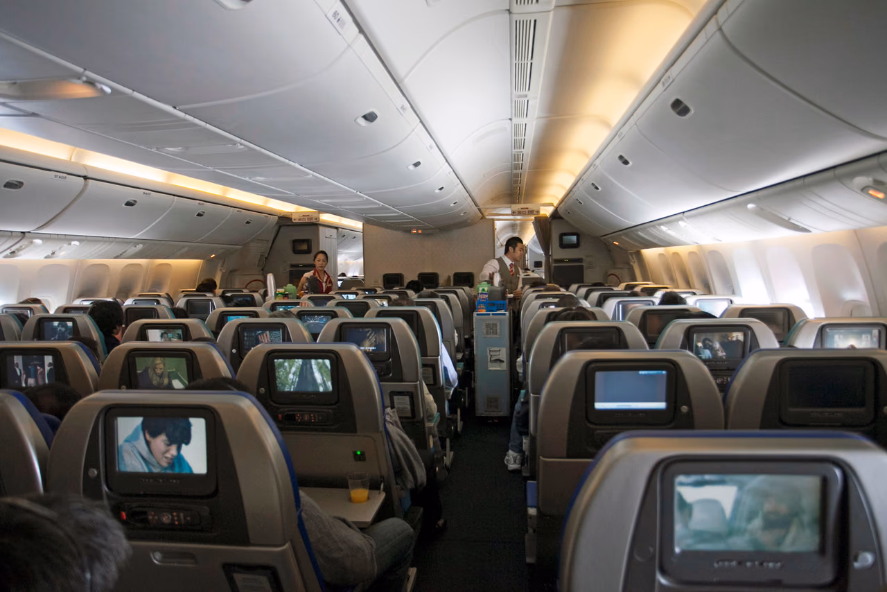 Flight attendants with a service cart in the aisle of a wide-body economy cabin, seen from behind rows of seats with seatback screens