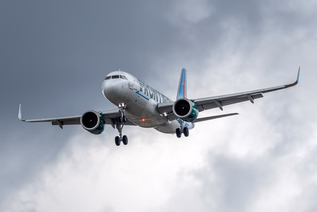 A Frontier Airlines Airbus A320neo on approach with its landing gear down, seen from below against heavy grey cloud, with the green Frontier titles visible along the forward fuselage