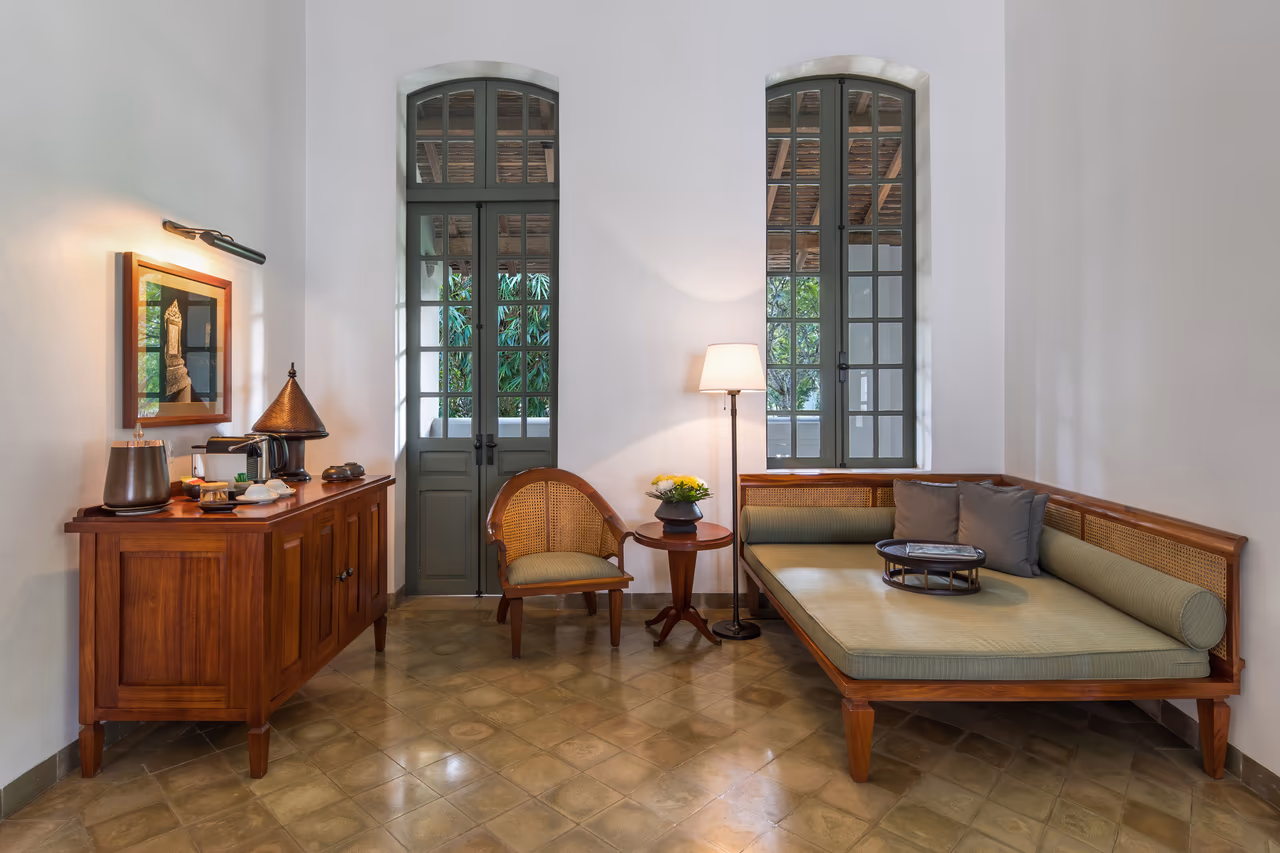 A hotel suite lounge with white walls and tall green-framed French doors opening onto greenery, a wooden daybed with pale green cushions, a caned armchair and a tea set on a wooden sideboard