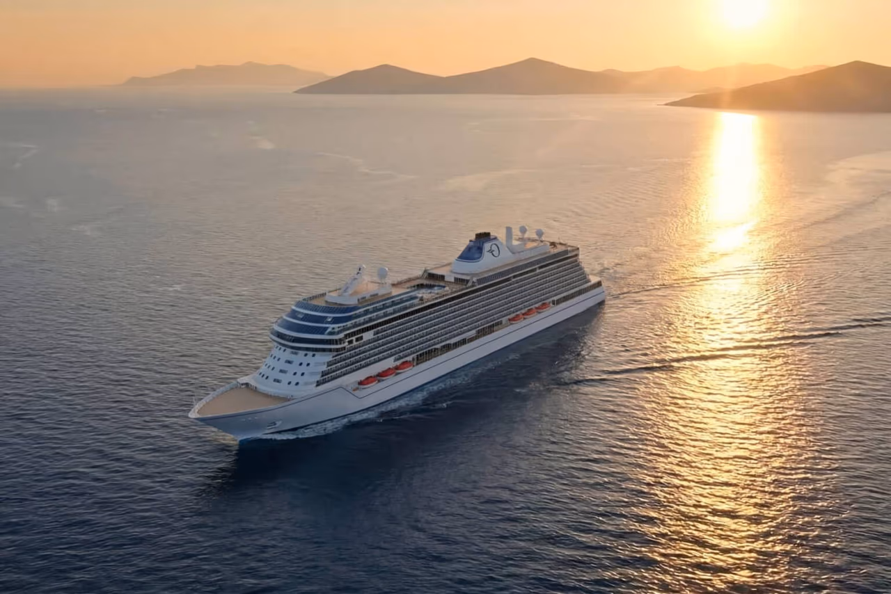 An aerial view of Oceania Sonata under way on calm open water at sunset, its white hull and tiered upper decks lit from the side, with low islands and a sun-path on the water behind