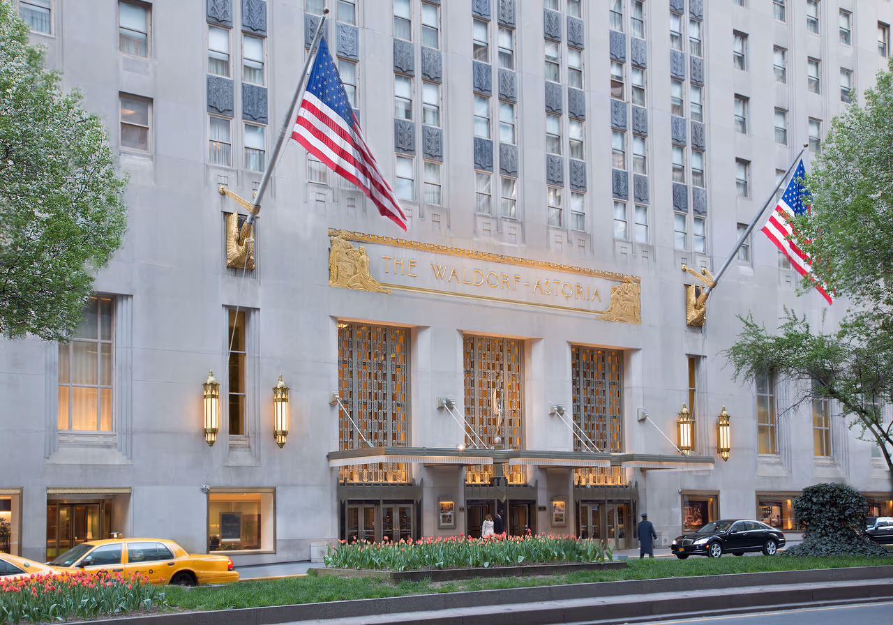 The Park Avenue entrance of the Waldorf Astoria in New York, its Art Deco limestone facade carrying gilded lettering above the doors, two American flags on angled poles, brass lanterns and tulip beds at the kerb