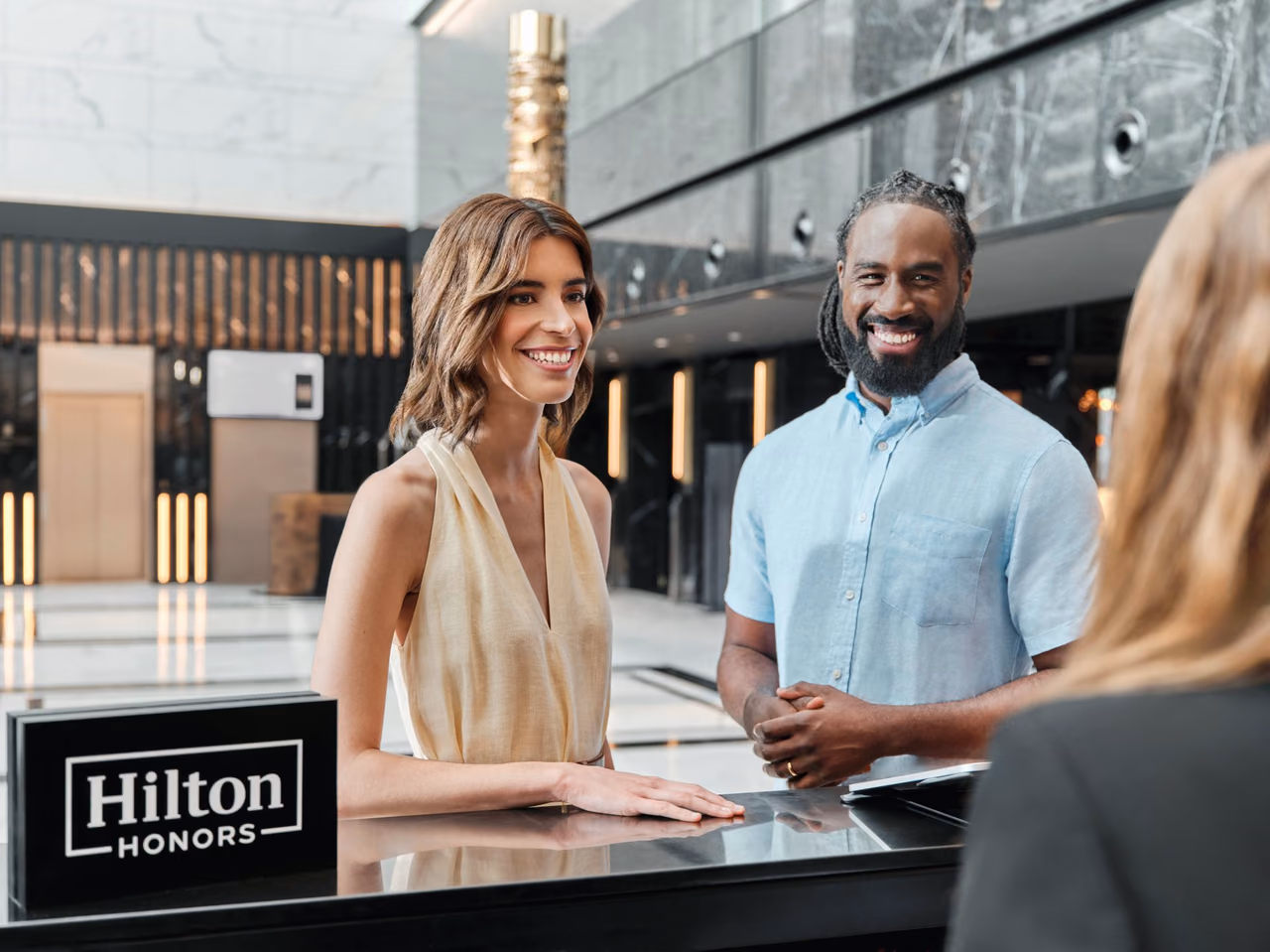 Two smiling guests at a dark, glossy hotel reception counter with a black Hilton Honors sign, talking with a front desk agent in a bright marble lobby