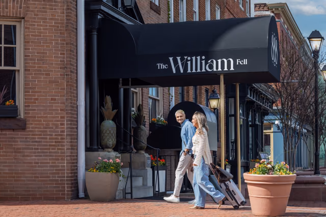 Two guests walking with a rolling suitcase past the entrance of The William Fell Baltimore, Tapestry Collection by Hilton