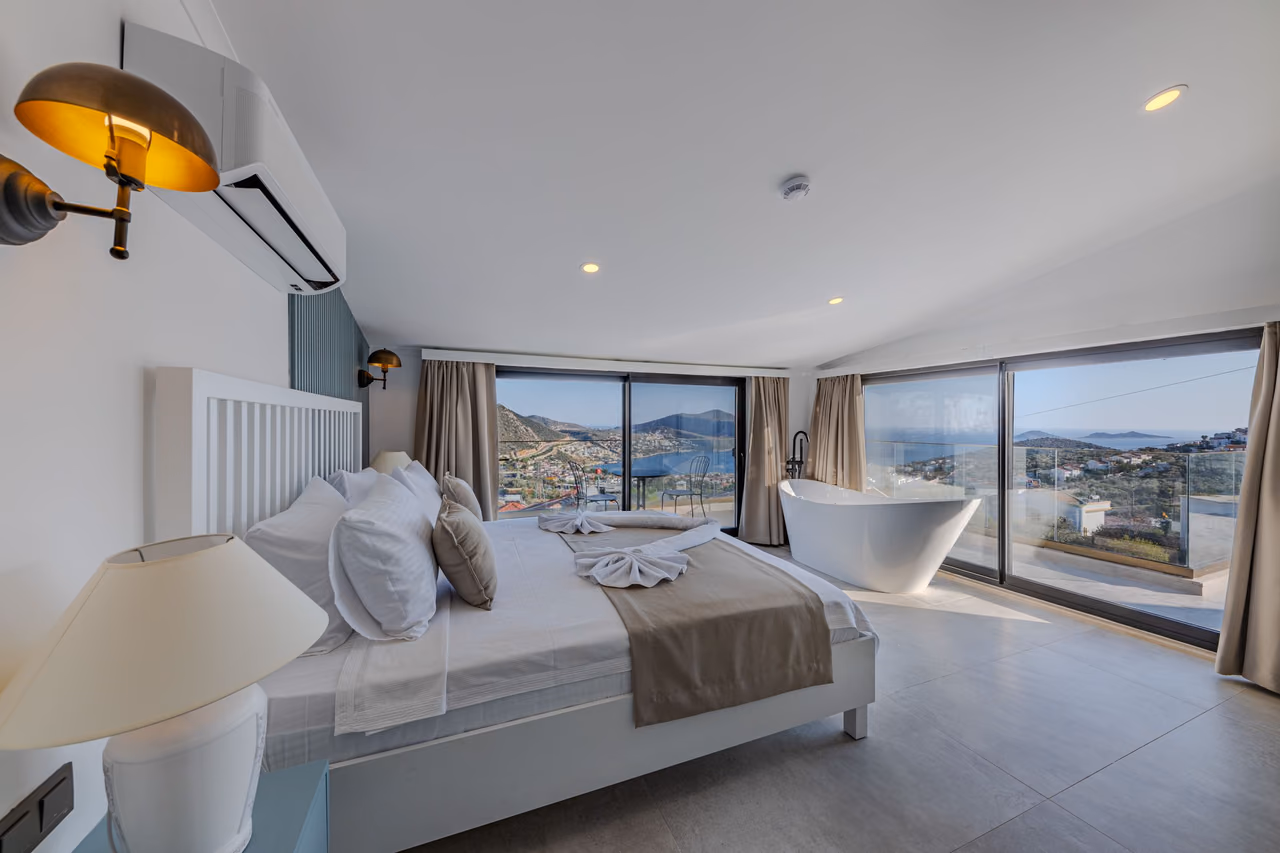 Hotel bedroom with a white bed and a freestanding bathtub beside floor-to-ceiling windows overlooking hills and the sea