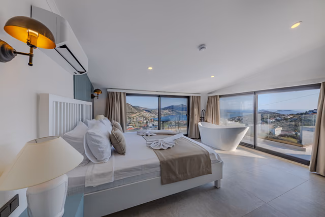 Hotel bedroom with a white bed and a freestanding bathtub beside floor-to-ceiling windows overlooking hills and the sea
