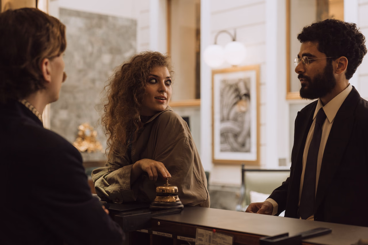 A guest with curly hair rests her hand beside a brass service bell at a hotel front desk while talking with a clerk in a dark suit and tie.