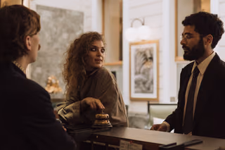 A guest with curly hair rests her hand beside a brass service bell at a hotel front desk while talking with a clerk in a dark suit and tie.