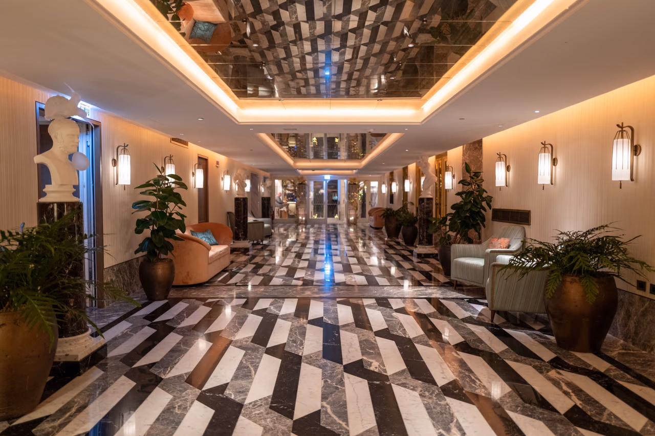 A luxury hotel hallway with a mirrored ceiling, striped marble floor, armchairs and potted plants.