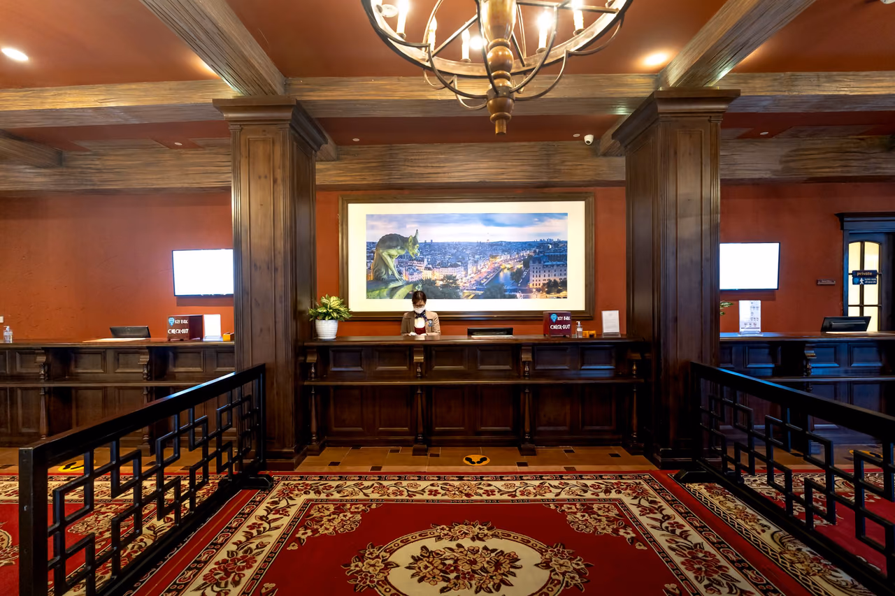 A hotel lobby with a wood-paneled front desk, a receptionist behind it, a wrought-iron chandelier overhead and a red patterned carpet