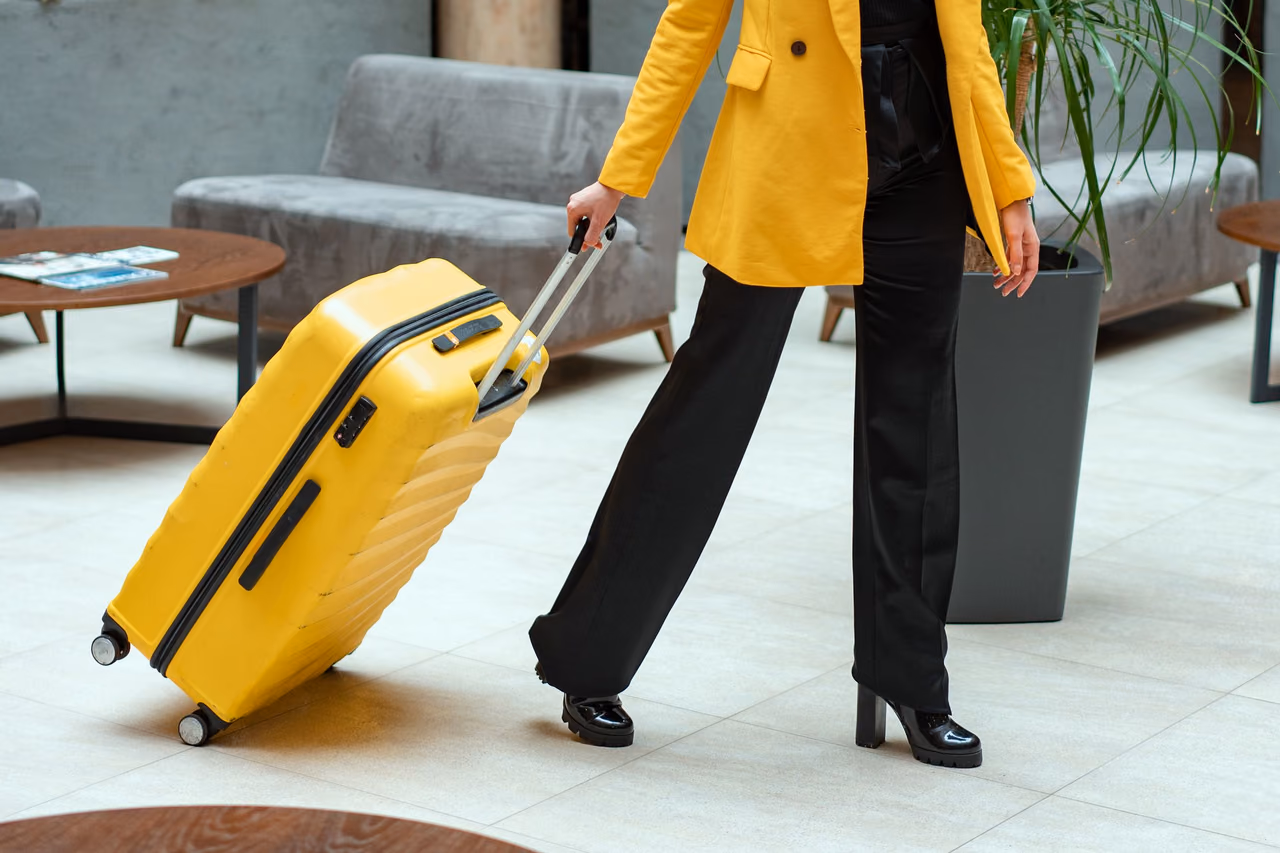 A traveler in a yellow blazer and black trousers pulling a yellow hard-shell suitcase across a hotel lobby with gray sofas