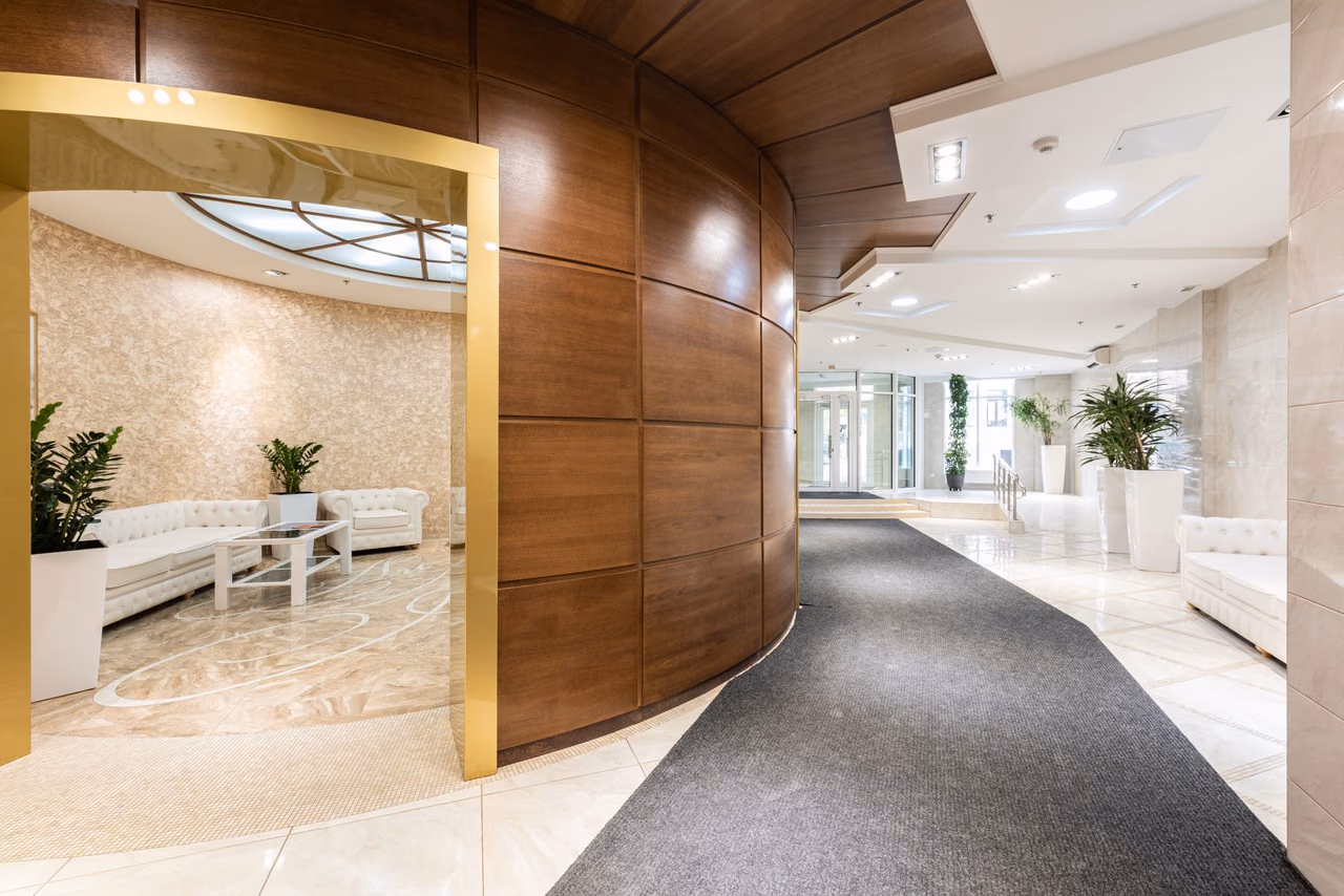 A bright hotel lobby with a curved wood-paneled wall, white sofas and potted plants beside a corridor of pale tile