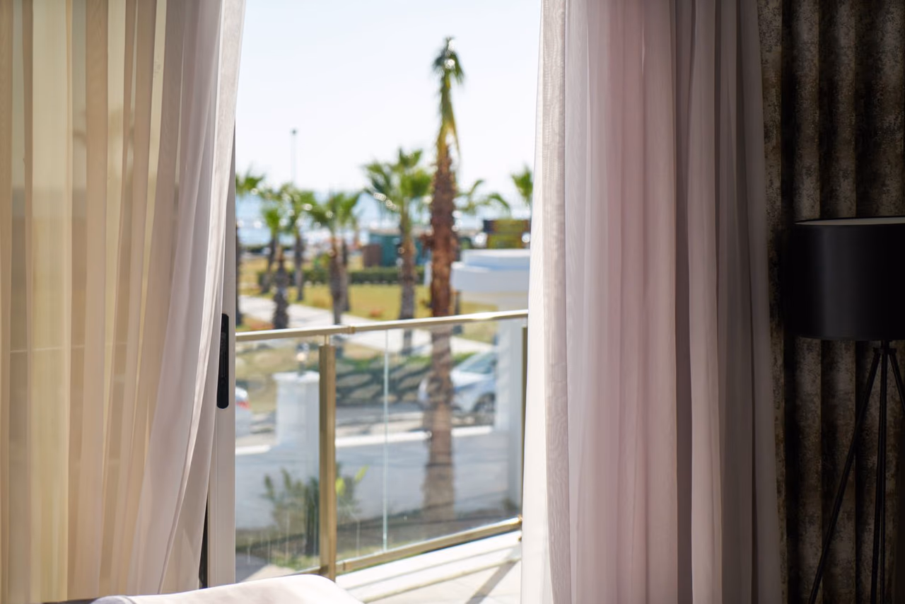 An open balcony door framed by sheer curtains, with a glass railing and palm trees outside a hotel room