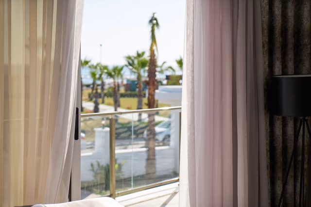 An open balcony door framed by sheer curtains, with a glass railing and palm trees outside a hotel room