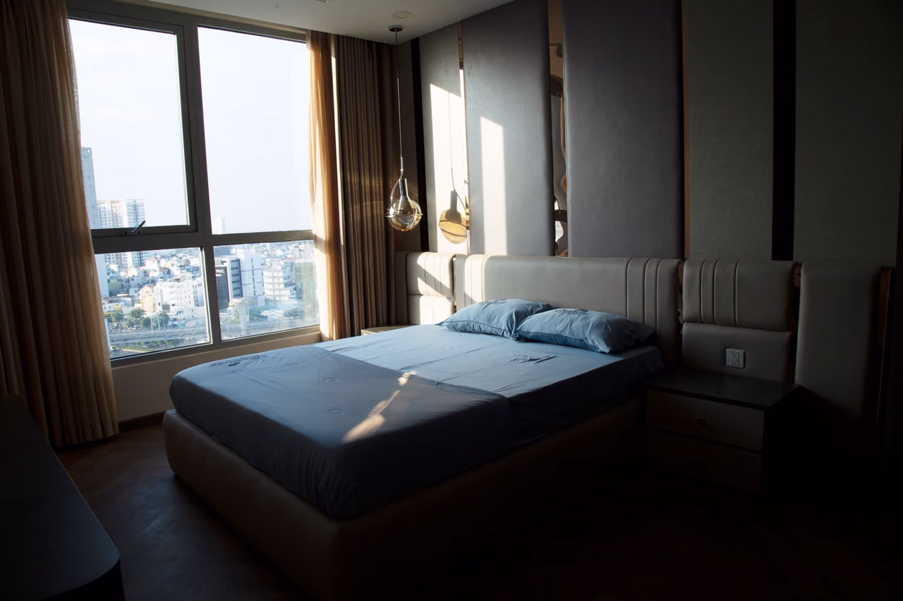 A hotel bedroom on a high floor with a double bed, blue linens and floor-to-ceiling windows looking out over a city in afternoon sun