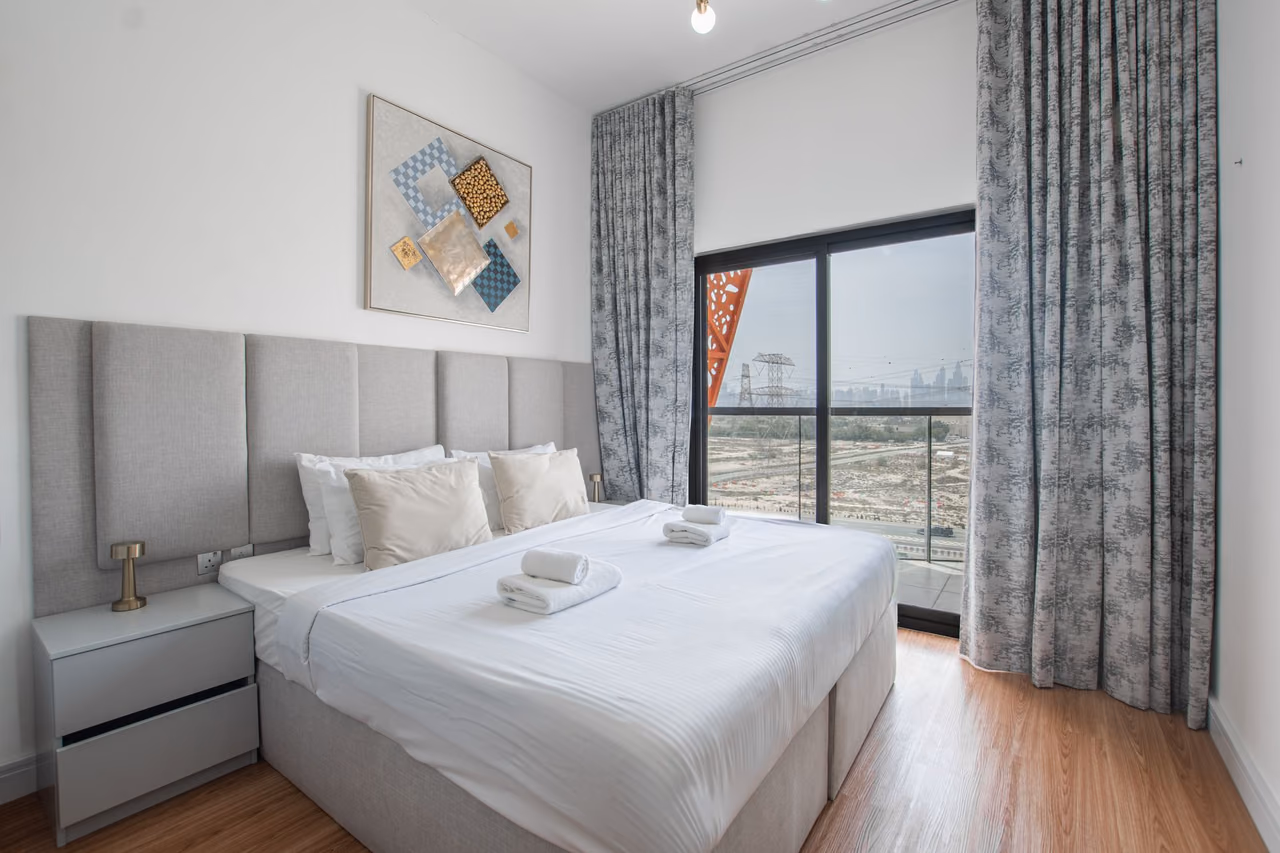 Freshly made hotel room with a king bed, folded towels, a gray padded headboard and a sliding glass door to a balcony