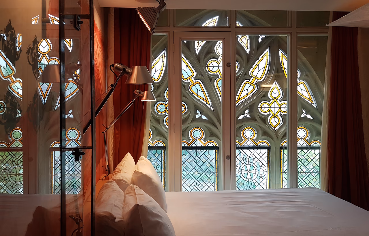 A hotel bed with white pillows in front of tall stained glass church windows, a reading lamp angled over the headboard