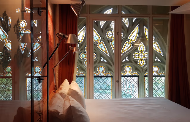 A hotel bed with white pillows in front of tall stained glass church windows, a reading lamp angled over the headboard