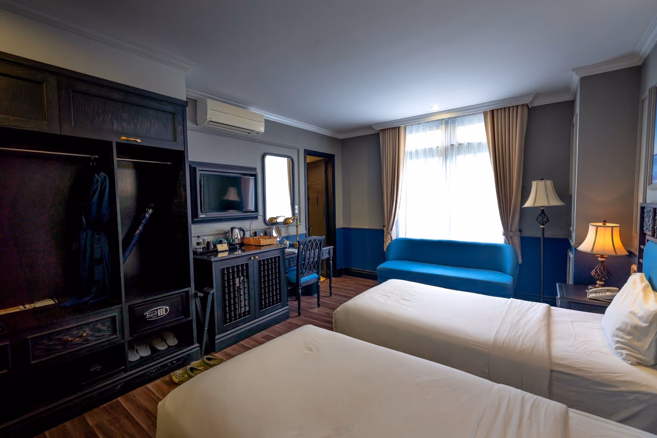 A bright hotel room with two made beds, a closet and natural light from the window
