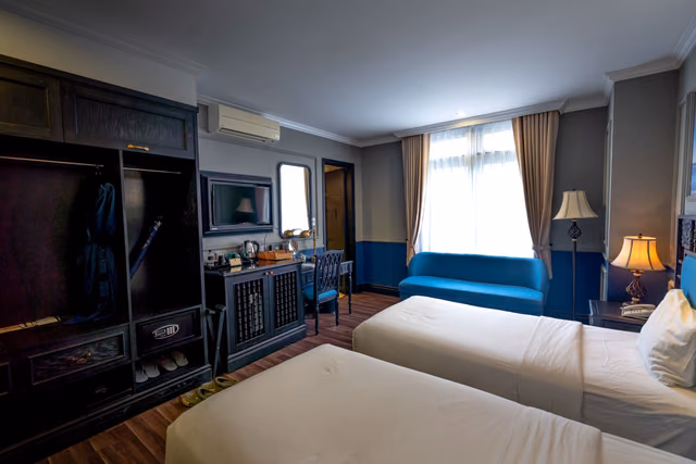 A bright hotel room with two made beds, a closet and natural light from the window