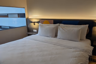 Hotel room with a made bed, four white pillows and a backlit headboard beside a window onto the next building