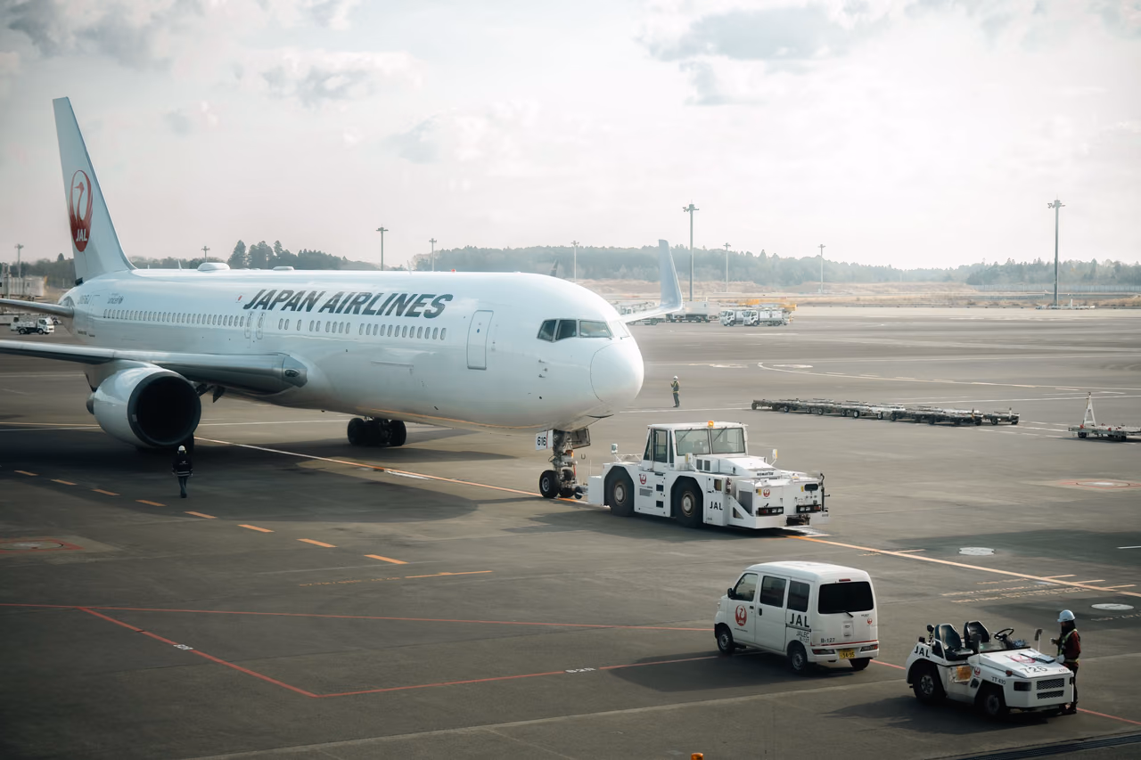 A Japan Airlines widebody jet with a red crane logo on its tail is pushed back by a tug on an airport apron, with ground service vehicles nearby.