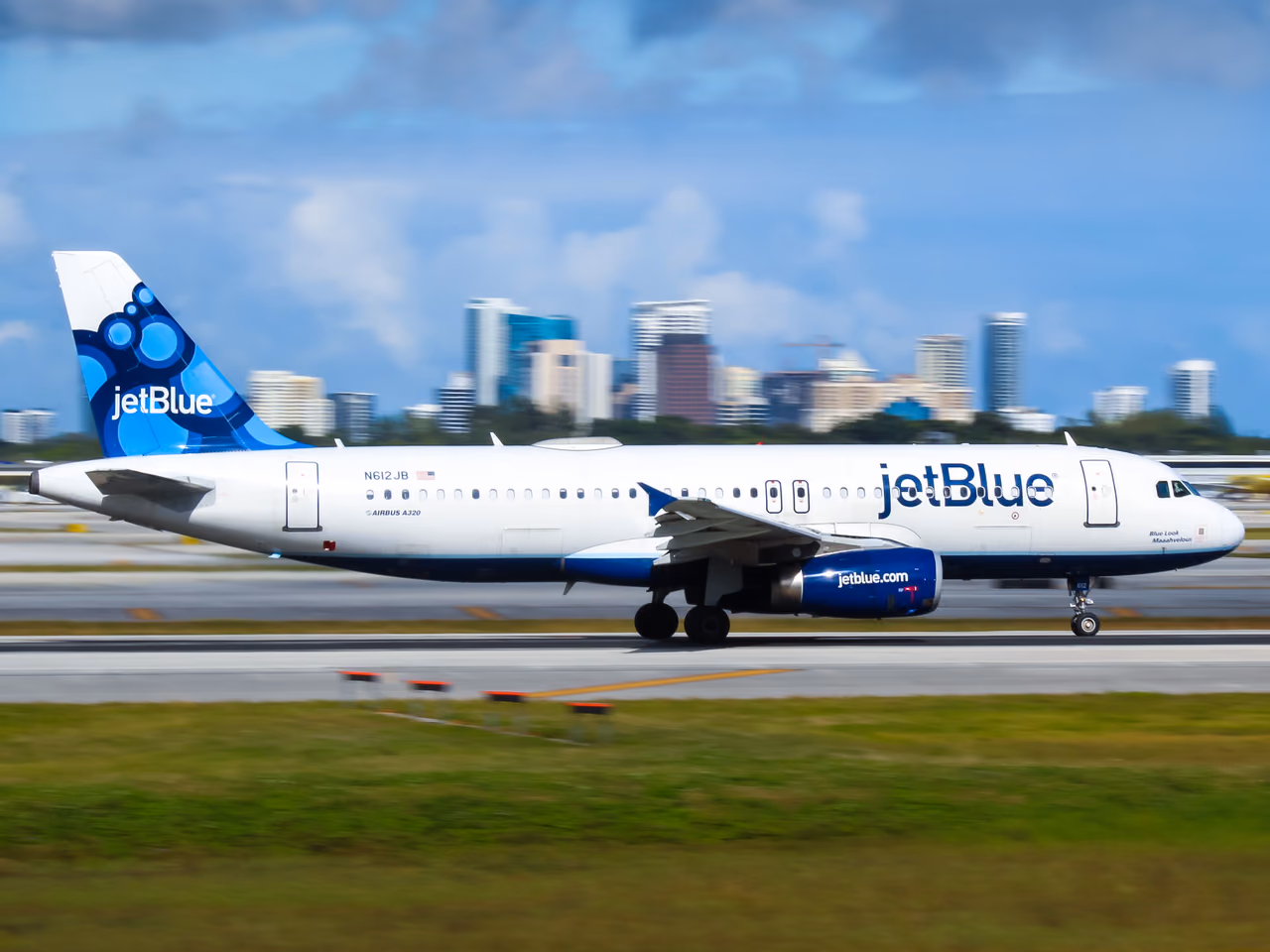 A JetBlue Airbus A320 rolling down a runway with a city skyline of high-rise towers behind it