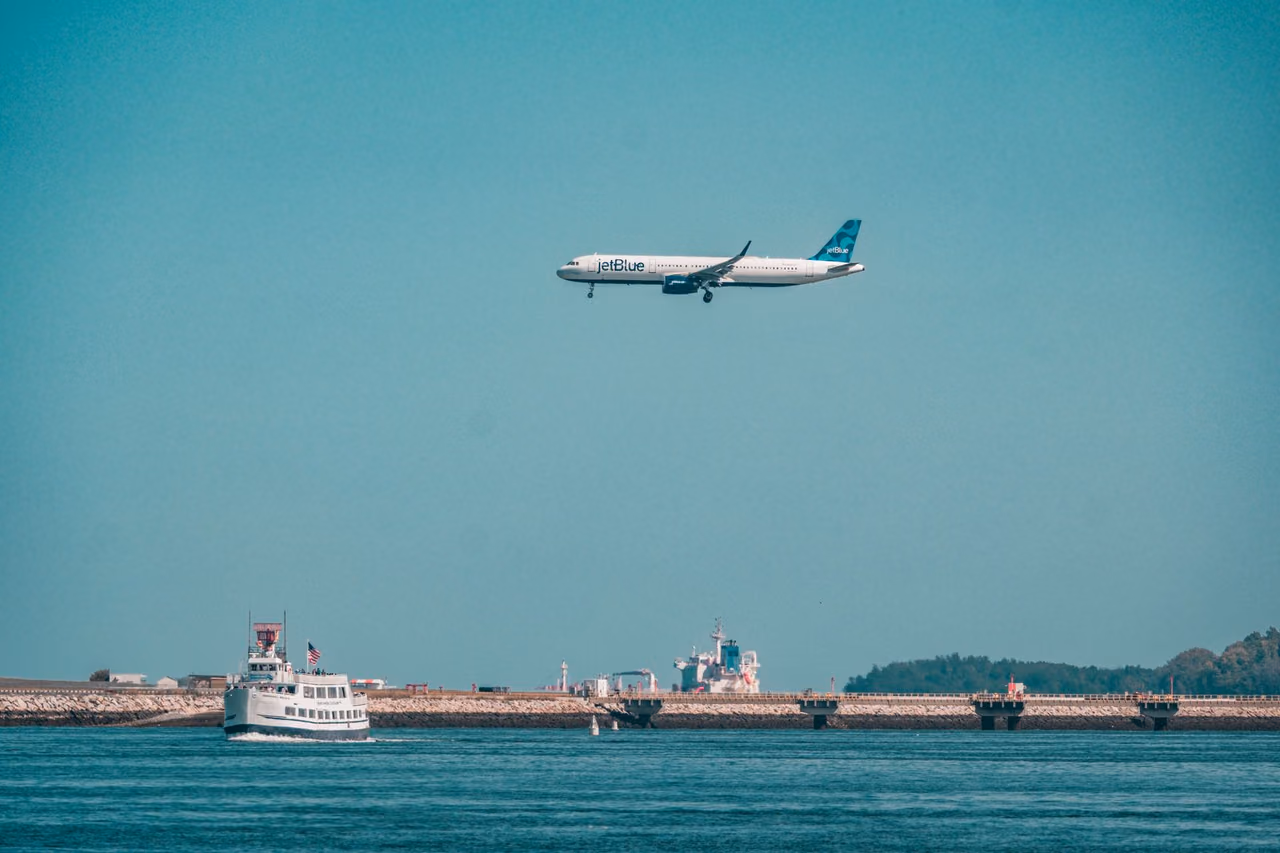 A JetBlue Airbus on approach over Boston Harbor, with a passenger ferry on the water below