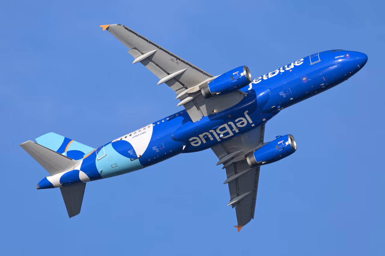 A JetBlue Airbus A320 in dark blue livery banking steeply after takeoff against a clear sky, landing gear retracted