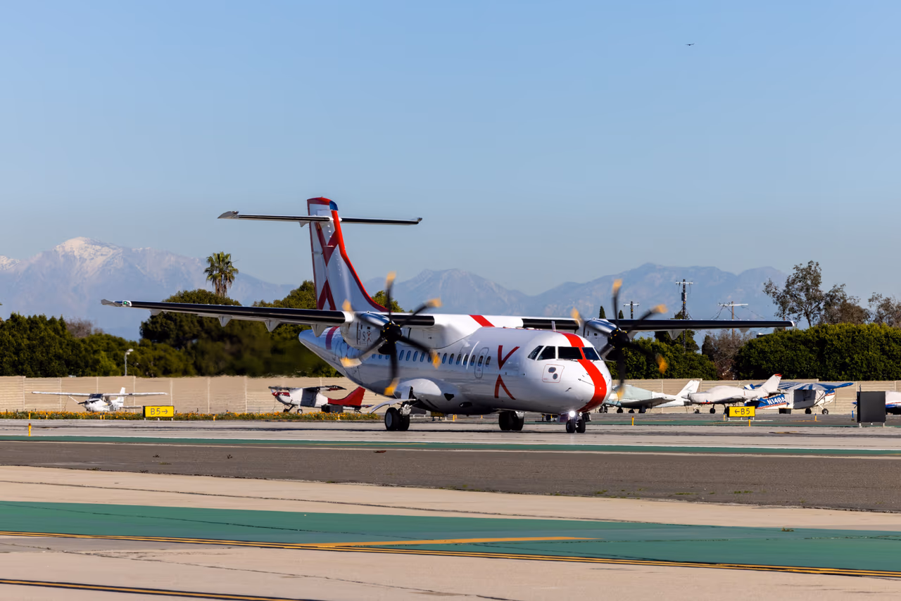 A JSX ATR 42-600 turboprop taxiing with its propellers turning, snow-capped mountains and parked light aircraft behind it