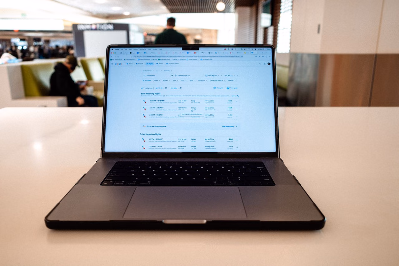 Open laptop on a white table in an airport lounge showing a flight search results page, with a traveler seated on a sofa behind it