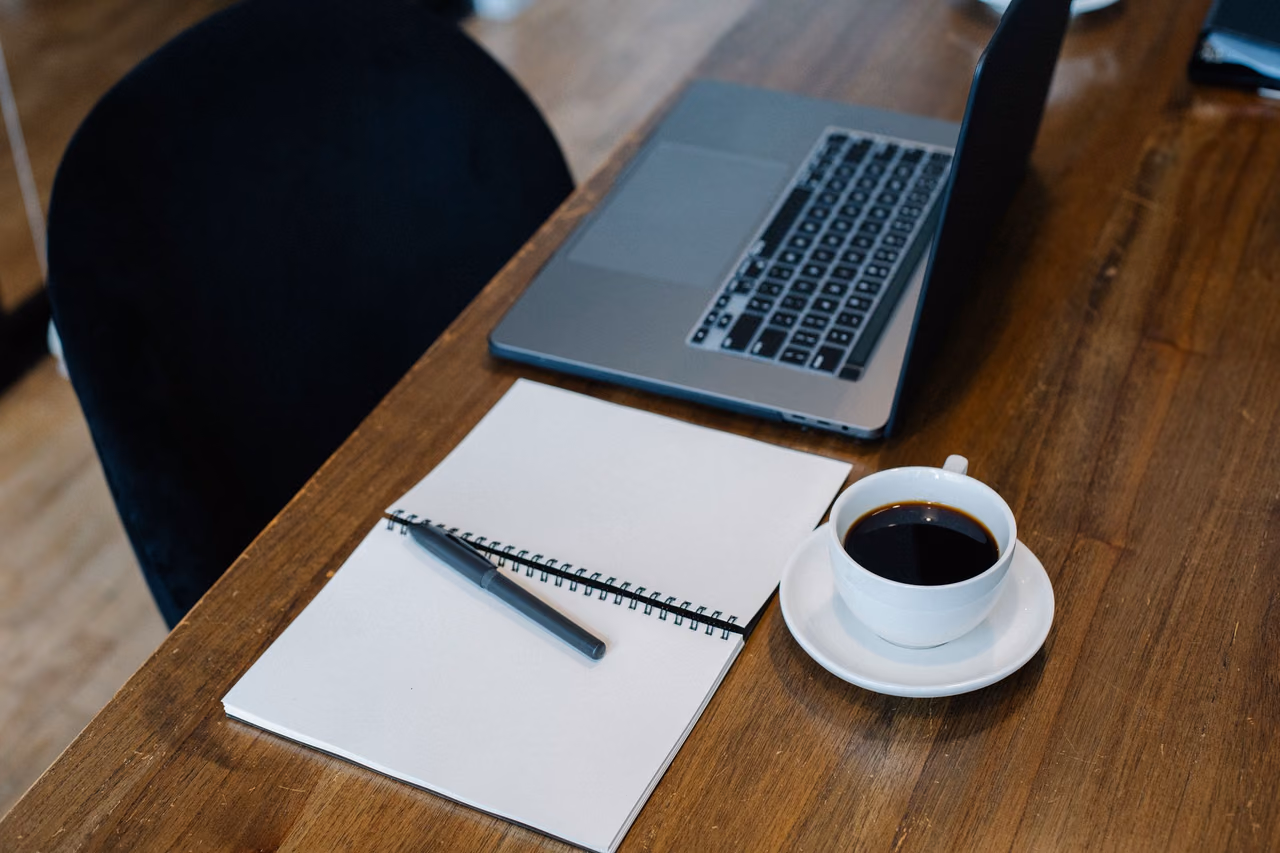 An open laptop, a spiral notebook with a pen and a cup of black coffee on a wooden desk.