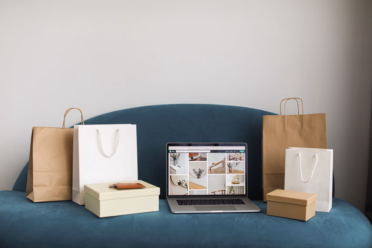 An open laptop on a teal sofa surrounded by paper shopping bags and gift boxes