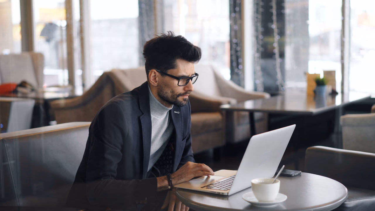A man in a blazer works on a laptop at a small cafe table with a cup of coffee beside him.