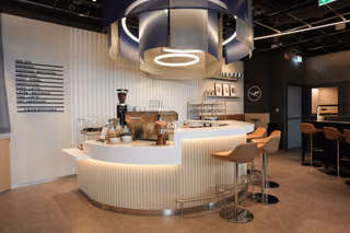 A curved barista coffee bar with tan stools in Lufthansa's renovated lounge at Stuttgart Airport, a coffee menu on the wall and the Lufthansa crane logo behind the counter