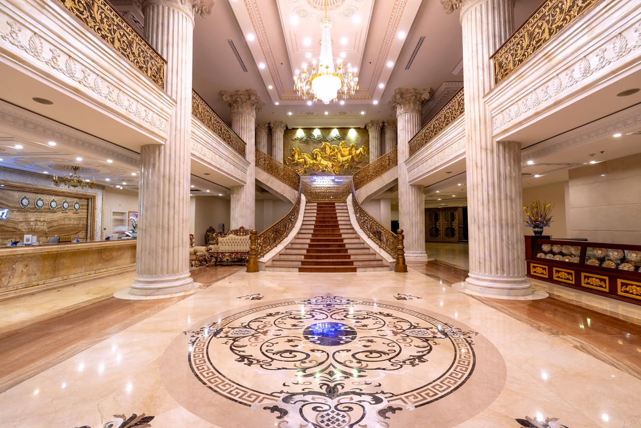 A marble hotel lobby with tall white columns, a chandelier and a red-carpeted grand staircase rising to a gilded mural.