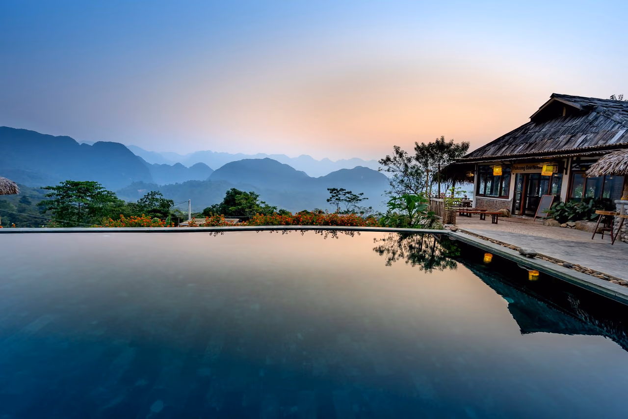 Swimming pool and terrace at a tropical resort with thatched buildings below a green mountain ridge