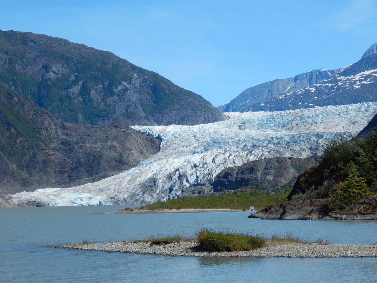 Mendenhall Glacier flowing between mountains into a pale blue lake near Juneau, Alaska, under a clear sky