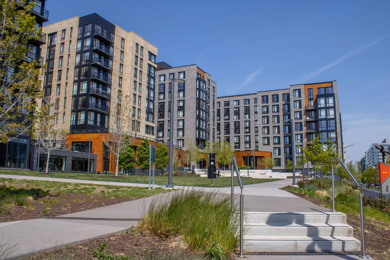 Modern brick and glass apartment buildings with balconies behind a landscaped plaza and concrete steps on a clear day.