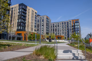 Modern brick and glass apartment buildings with balconies behind a landscaped plaza and concrete steps on a clear day.