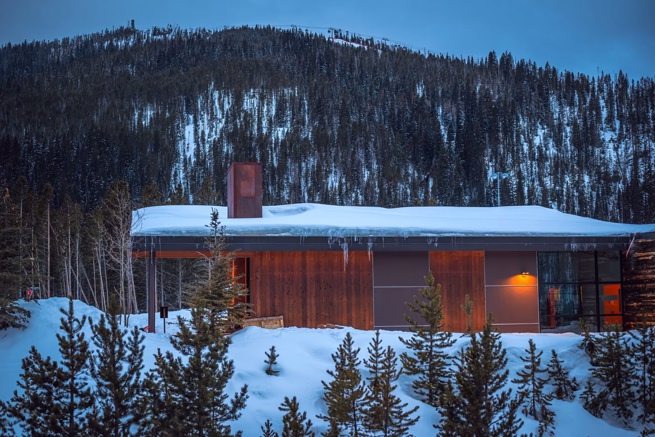 A modern wood-clad cabin with a snow-covered roof and a glowing window, set among snowy pines below a forested mountain at twilight
