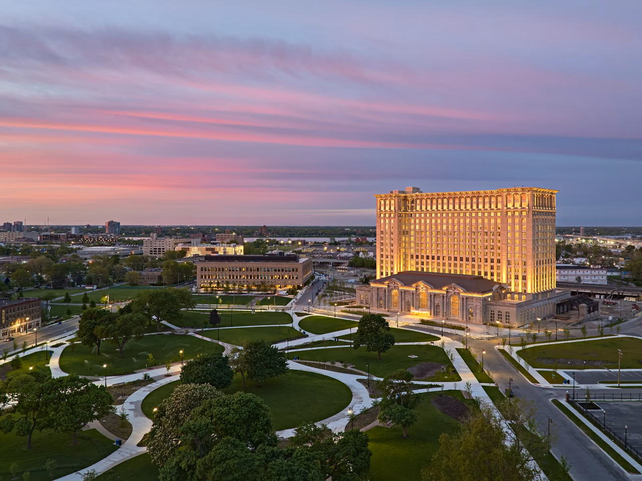Michigan Central Station's 15-story tower lit gold at dusk, with its low base and landscaped park paths below under a pink sky