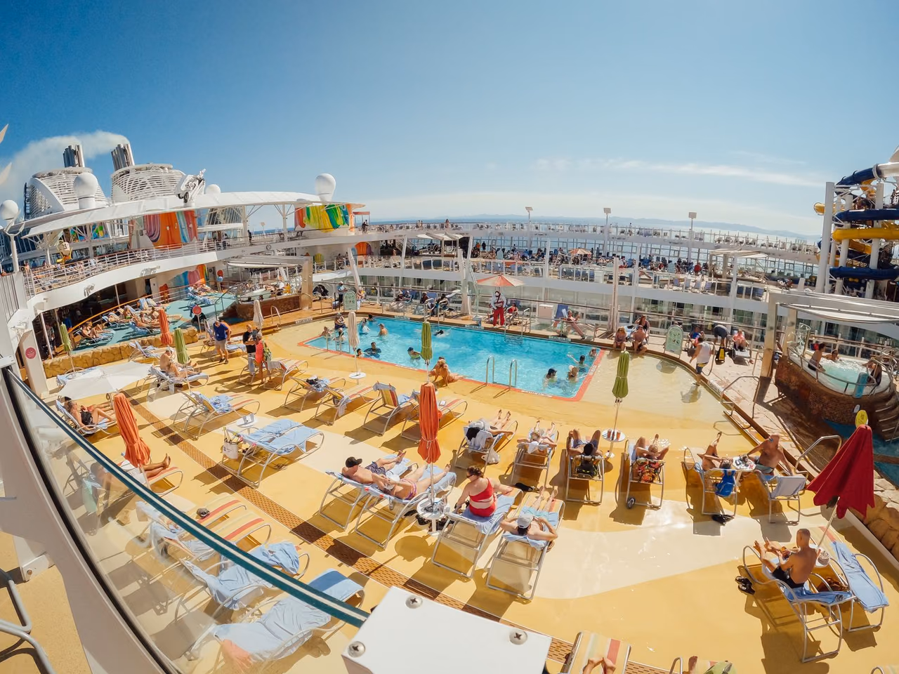 Passengers relax on loungers around the main pool on the pool deck of Royal Caribbean's Oasis of the Seas, with waterslides on the right and the open sea beyond