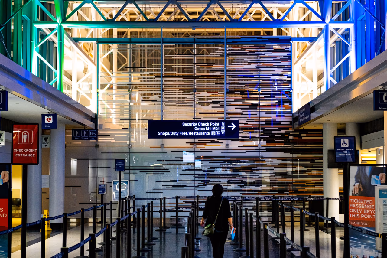 A traveler walks through the queue lines toward an airport security checkpoint entrance beneath a lit glass atrium