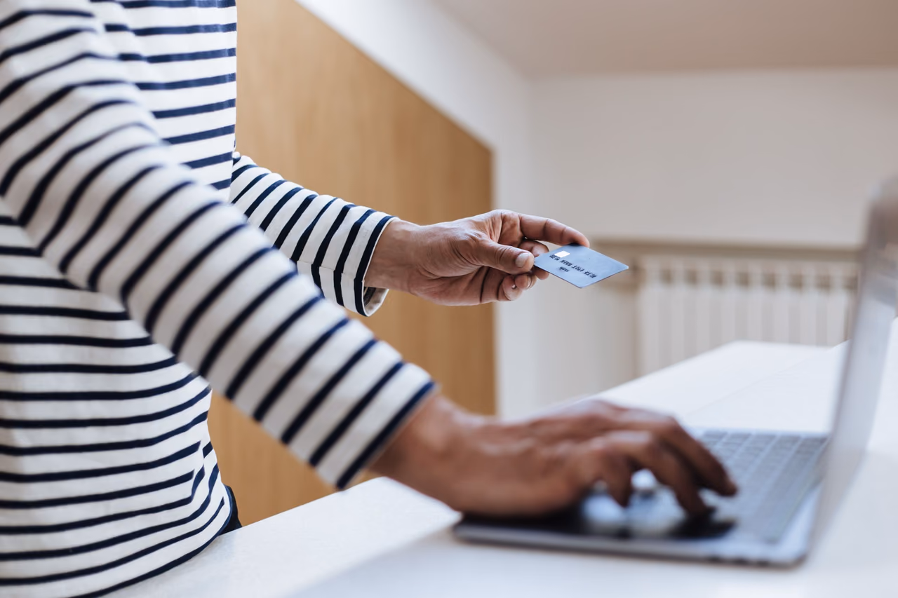 A person in a striped shirt holds a blue payment card in one hand and types on a laptop with the other at a white counter