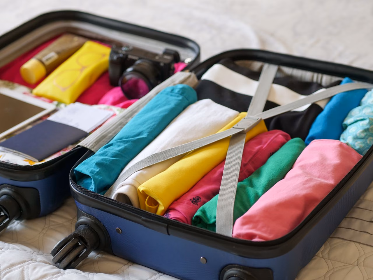 An open blue carry-on suitcase packed with rolled colorful shirts under a cross strap, with a camera, sunglasses case and passport in the lid