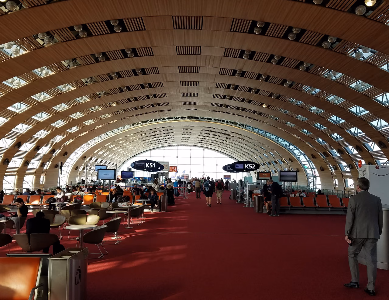 Gate area at Paris Charles de Gaulle Terminal 2E under a curved wood-slat roof, with red carpet, orange seats and signs for gates K51 and K52