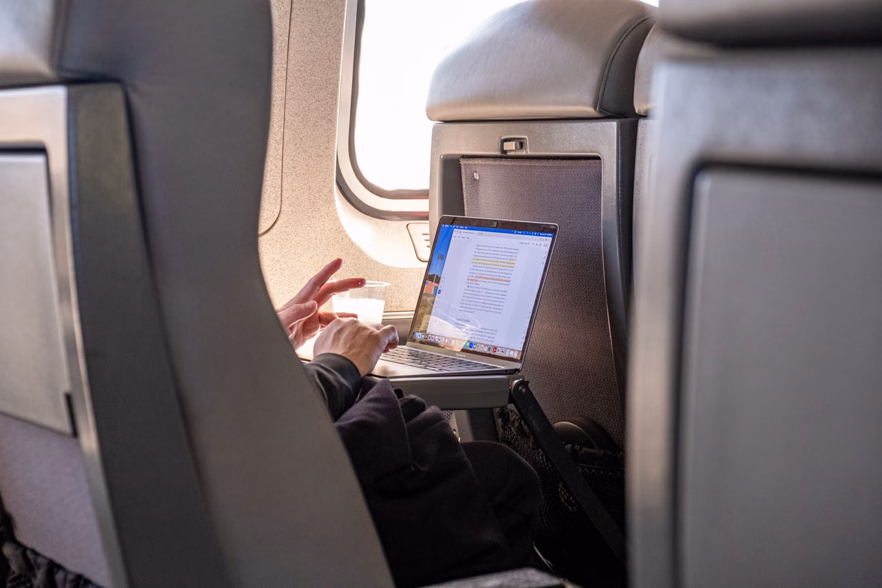 A passenger typing on a laptop on a seatback tray table beside an airplane window during a flight