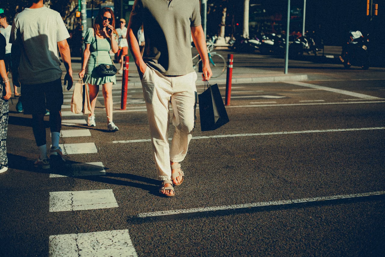 Pedestrians crossing a sunlit city street, with a man in the foreground carrying a black paper shopping bag and a woman behind him holding a tote.